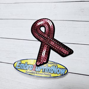 May include: Pink sequined ribbon patch with the text "Embro & PrintNow Embroidery & Screen Printing" on a yellow oval sticker.