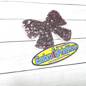 May include: A pink sequined bow applique with a white backing. The applique is on a white wooden surface. A yellow oval sticker with the text "Embro dPrintNow Minneapolis Embroidery & Screen Printing" is in the lower right corner of the image.