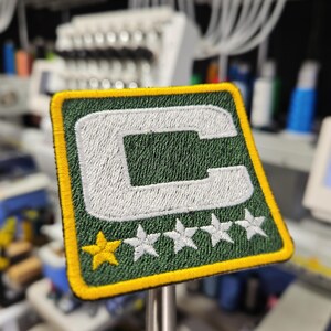 May include: Embroidered patch with a green background, a white letter "C", and five white stars. The patch has a yellow border and a gold star. The patch is square and is likely for a sports team or organization.