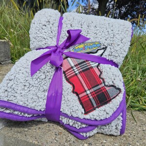 May include: A gray sherpa blanket with a purple trim and a purple ribbon tied around it. The blanket has a plaid patch with red, black, and white colors. The blanket is folded and tied with a purple ribbon.