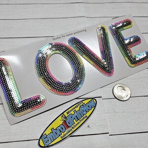 May include: A close-up of a white card with the word "LOVE" spelled out in large, colorful, sequined letters. The letters are rainbow-colored and have a silver glitter finish. The card also has the text "Sticker for easier positioning" and a small business card that says "Embro dPrintNow Minneapolis Embroidery & Screen Printing".