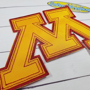 May include: Embroidered patches featuring the letter "M" in gold with a maroon outline. The patches are on a white wooden surface. The patches are likely for apparel or accessories.