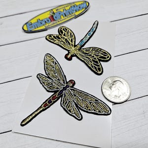 May include: Two gold and silver embroidered dragonfly iron-on patches. The dragonflies have a detailed design with black outlines and blue accents.