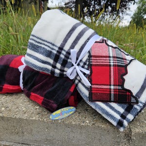 May include: Folded blankets with plaid patterns in red, black, and white, tied with white ribbons. One blanket features a red and black plaid design, while another has a white and gray striped pattern. The blankets are stacked on a concrete surface.