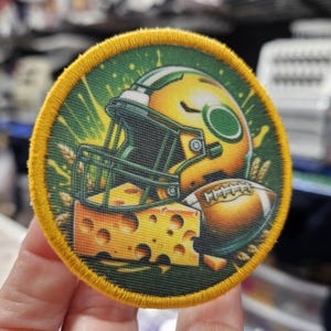 May include: Circular embroidered patch featuring a football helmet, a football, and a block of cheese. The patch has a green background with yellow accents and a yellow border. The design is likely related to a sports team.