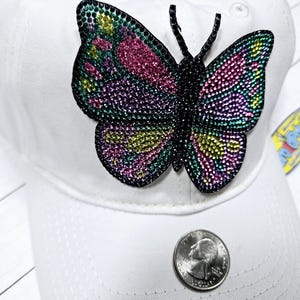 May include: White baseball cap with a large, colorful butterfly rhinestone applique. The butterfly has pink, green, and yellow rhinestones.