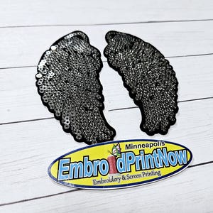 May include: A pair of black angel wings made of silver sequins. The wings are on a white wooden surface. The text "EmbroiderPrintNow" is visible in the lower right corner of the image.