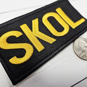 May include: Black rectangular patch with the word "SKOL" embroidered in yellow thread. A quarter is next to the patch for size comparison. The patch has a textured surface.