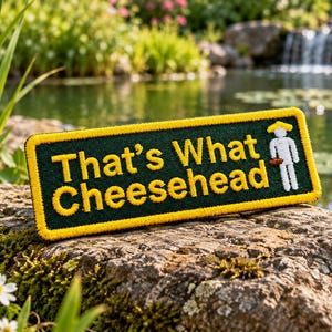 May include: A rectangular embroidered patch with a dark green background and a yellow border. The text "That's What Cheesehead" is stitched in yellow, accompanied by a small white stick figure wearing a yellow hat and holding a brown object.