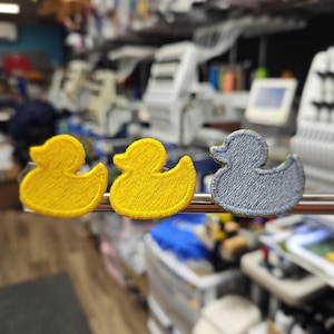 May include: Three embroidered duck patches. Two are yellow, and one is gray. The ducks are displayed on a metal bar in front of a blurred background of sewing machines and shelves.