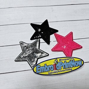 May include: Three sparkly star-shaped iron-on patches in black, silver, and pink. The patches are on a white wooden surface. The text "Embroider & Print Now" is visible in the lower right corner of the image.