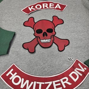 May include: Gray hoodie with green sleeves and hood lining. Features a red skull and crossbones patch, a red patch that says "KOREA", and a red patch that says "HOWITZER DIV." in white letters.