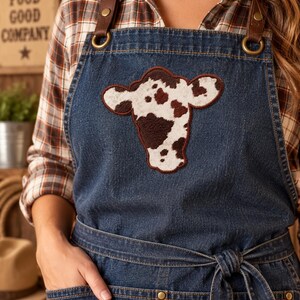 May include: A dark blue denim apron featuring a central cow head patch with a white and brown spotted pattern. The apron includes brown leather shoulder straps attached with brass-colored metal rings and rivets, plus a front waist tie and pockets.