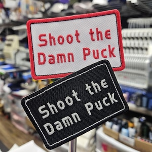 May include: Two embroidered patches with the text "Shoot the Damn Puck." One patch is rectangular with a white background and red border, the other is black with a white border. Both patches are on a metal rod.