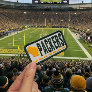 May include: A rectangular embroidered patch featuring a white background with a dark green border. The patch displays the word PACKERS in dark green capital letters, accompanied by a yellow silhouette of the state of Wisconsin and yellow stitching.