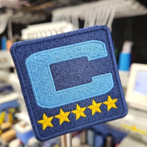 May include: Embroidered patch with a navy blue background and a light blue letter "C". The patch has five yellow stars at the bottom. The patch is square and has a navy blue border.