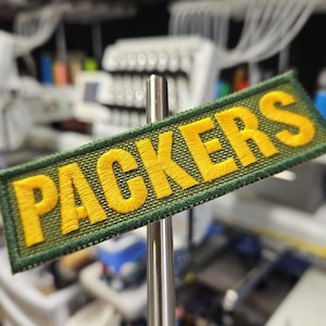 May include: A rectangular embroidered patch with the word "PACKERS" in yellow, stitched onto a green background. The patch is held by a metal rod, with a blurred workshop background.