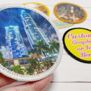 May include: A white, round, fabric patch with a cityscape image of tall buildings and palm trees. The patch has a white stitched border. The text "Custom Graphic or Text Here!" is on a yellow background with a black border.