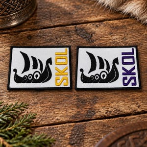 May include: Two rectangular embroidered patches with a white background and a black border. Each patch features a black Viking ship silhouette and the word SKOL embroidered in either yellow or purple thread on the right side of the design.