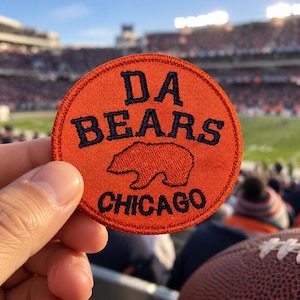 May include: A circular orange embroidered patch featuring the dark blue text DA BEARS and CHICAGO with a dark blue bear silhouette in the center. The patch has a stitched border and is held against a blurred background of a football stadium.