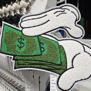 May include: An embroidered patch featuring two white cartoon-style gloved hands holding a stack of green paper money with a black dollar sign symbol. The patch has a thick white border and is positioned in front of an industrial embroidery machine.
