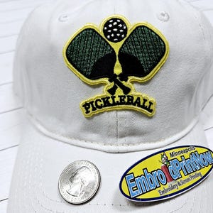 May include: White baseball cap with a yellow and black embroidered patch that says "Pickleball" and depicts two pickleball paddles and a ball.