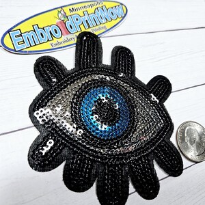 May include: A black and blue evil eye patch with sequins. The eye is surrounded by silver sequins and the pupil is black. The patch is shaped like a hand with five fingers.