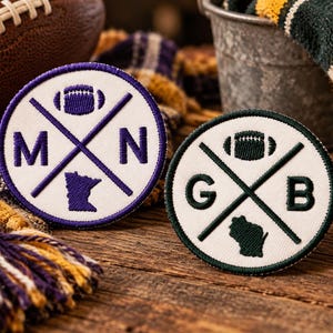 May include: Two circular embroidered patches with white backgrounds. The left patch features purple stitching, the letters M and N, a football, and the state of Minnesota. The right patch features dark green stitching, the letters G and B, a football, and the state of Wisconsin.