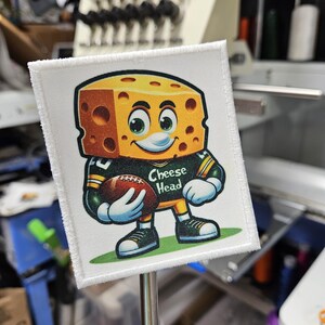 May include: A square patch featuring a cartoon cheese block character in a football uniform, holding a football. The patch has a white border and the text "Cheese Head". The character is yellow with brown holes, wearing a green and gold uniform.