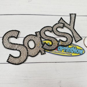 May include: Close-up of a rhinestone iron-on patch that says "SASSY" in black outlined letters. The patch is on a white wood background.