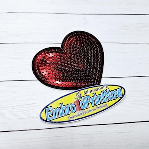 May include: Red heart-shaped sequin patch with black edging. The patch is on a white wooden surface. The text "Embro & PrintNow" is on a blue and yellow oval sticker with the text "Minneapolis" and "Embroidery & Screen Printing".