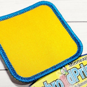 Könnte beinhalten: Ein quadratischer gelber Patch mit einem blauen Rand. Der Patch ist aus gewebtem Stoff und hat einen genähten Rand.