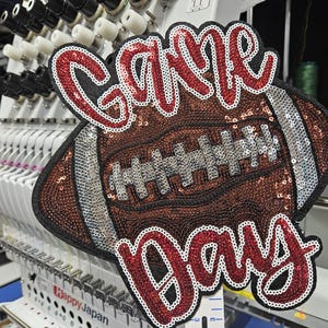May include: A sequined patch featuring a brown football with silver laces and the text Game Day in red script. The patch is outlined in black and is positioned in front of an embroidery machine. The design uses red, brown, silver, and black sequins.