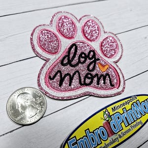May include: A pink glitter dog paw patch with the words "dog mom" embroidered in black script. The patch has a white border and an orange heart accent. A quarter is next to the patch.