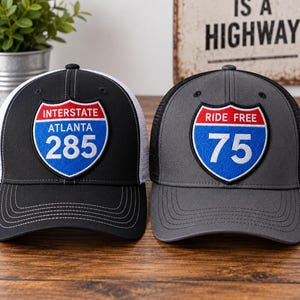 May include: Two baseball caps with mesh backs. The black cap features an embroidered patch reading INTERSTATE ATLANTA 285. The grey cap features an embroidered patch reading RIDE FREE 75. Both patches are shield-shaped with red, white, and blue colors.