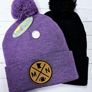 May include: Two knit beanies with pom poms. One beanie is purple with a wooden patch that says "MN" with a football and the outline of Minnesota. The other beanie is black.