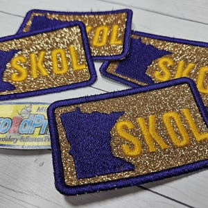May include: Embroidered patches with a gold glitter background, a purple outline of the state of Minnesota, and the word "SKOL" in gold. The patches are rectangular with rounded corners.