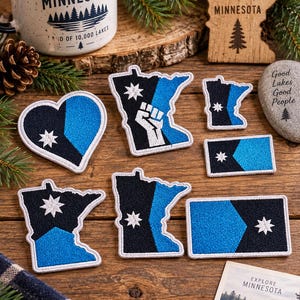 May include: A collection of embroidered patches featuring the Minnesota state flag design in black, blue, and white. The set includes various shapes such as hearts, rectangles, and the outline of the state of Minnesota, some with a white fist icon.
