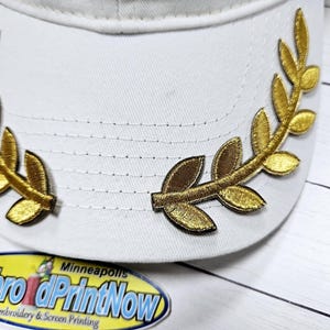 May include: A white baseball cap with two gold embroidered laurel wreaths on the front. The cap is on a white wooden surface.