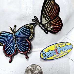 May include: Two embroidered butterfly patches on a white baseball cap. The butterflies are both detailed with colorful thread and have a black outline. The larger butterfly is blue, red, and gold. The smaller butterfly is orange, yellow, and gold. The cap also has a yellow oval sticker with the text "Embro & Print Now" in black.