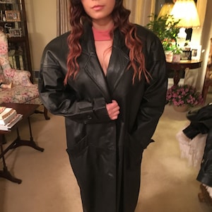 May include: A long black leather coat with a button closure and pockets. The coat is worn by a person with long brown hair.
