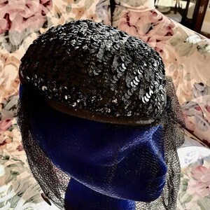 May include: A vintage black sequin hat with a net veil. The hat is oval-shaped and covered in small, shiny black sequins. A black net veil drapes over the front. The hat is displayed on a blue mannequin head.