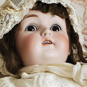 May include: A porcelain doll with brown hair wearing a white lace bonnet and a white dress with lace trim.