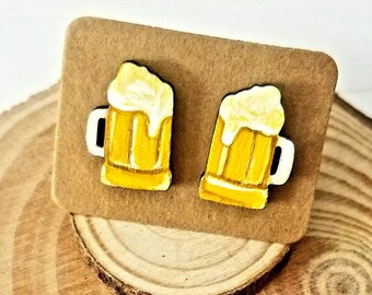 May include: A pair of small, yellow beer mug earrings with white foam on top. The earrings are on a brown card and a wooden coaster with a penny on top.