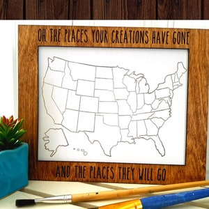 May include: A wooden frame with a white outline of the United States map. The frame has the text "Oh the places your creations have gone and the places they will go" printed on it. Two paintbrushes are in the foreground.