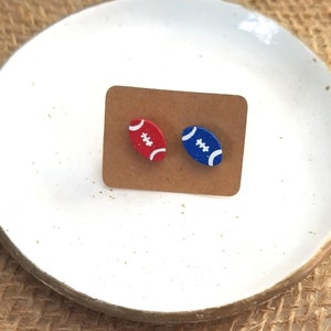 May include: A pair of football-shaped earrings on a brown card. One earring is red with white stitching and the other is blue with white stitching.