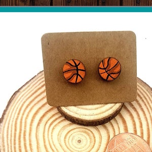 May include: Hand painted basketball earrings on a wooden display stand. The earrings are orange and black with a white outline. The stand is made of wood and has a natural finish. The text "Hand Painted Earrings" is written in blue on a blue watercolor background.