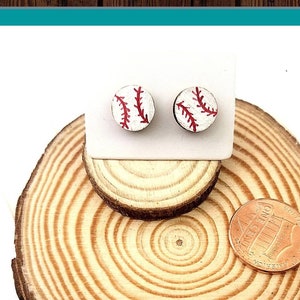 May include: A pair of hand-painted baseball earrings on a wooden display stand. The earrings are white with red stitching and are displayed on a wooden round with a coin for size reference. The text "Hand Painted Earrings" is visible in the image.