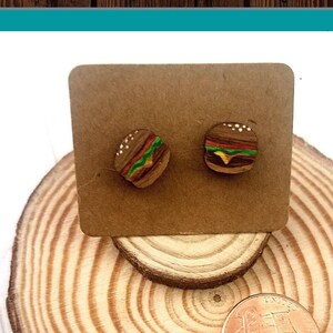 May include: Hand painted hamburger earrings on a wooden display stand. The earrings are small and detailed, with a brown bun, yellow cheese, green lettuce, and red tomato. The stand is made of wood and has a circular shape. The text "Hand Painted Earrings" is written in blue on a white background.