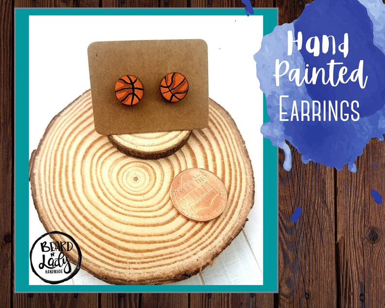 May include: Hand painted basketball earrings on a wooden display stand. The earrings are orange and black with a white outline. The stand is made of wood and has a natural finish. The text "Hand Painted Earrings" is written in blue on a blue watercolor background.